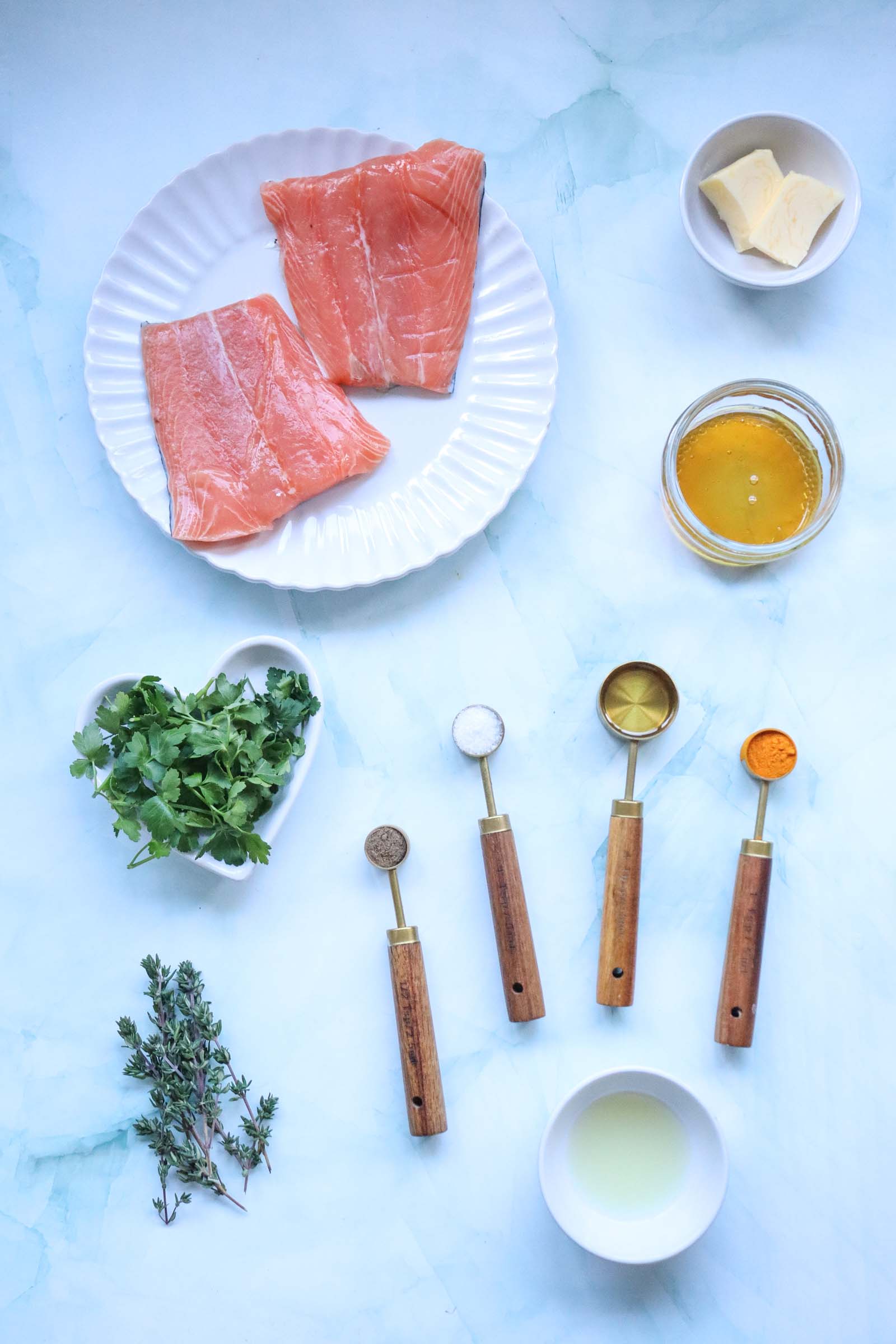 Overhead view of raw salmon fillets on a plate, honey, butter, fresh herbs, and measured spices on a light blue background.