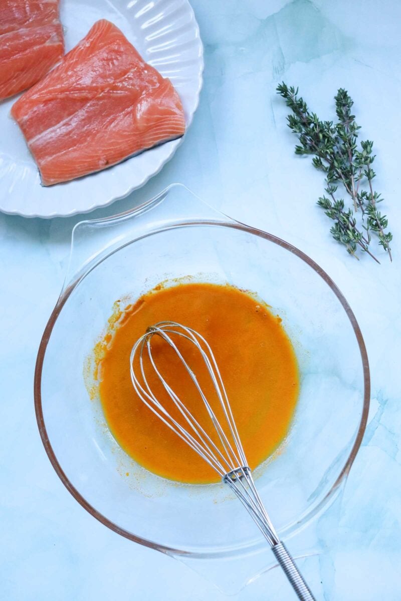 A bowl of glaze with a whisk, fresh salmon fillets on a plate, and sprigs of thyme on a light blue surface.