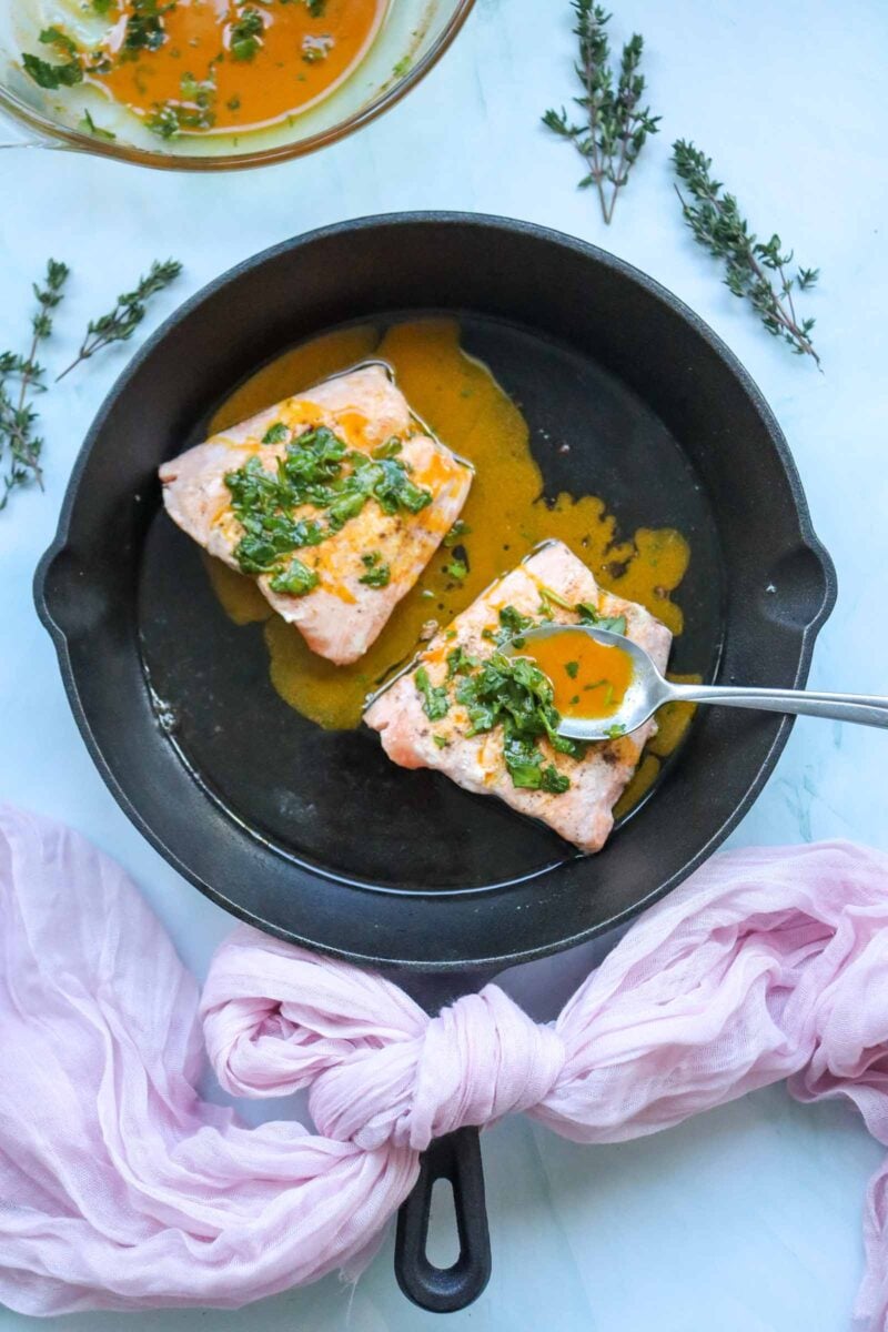 Two cooked salmon fillets in a black skillet, topped with green herbs and honey glaze, with a spoon resting on one fillet; fresh herbs and a pink cloth are nearby.