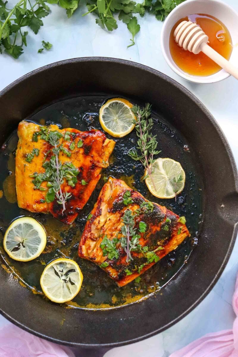 Two pieces of cooked honey glazed salmon in a cast iron skillet, garnished with lemon slices, fresh herbs, and a honey dipper in a bowl of honey nearby.