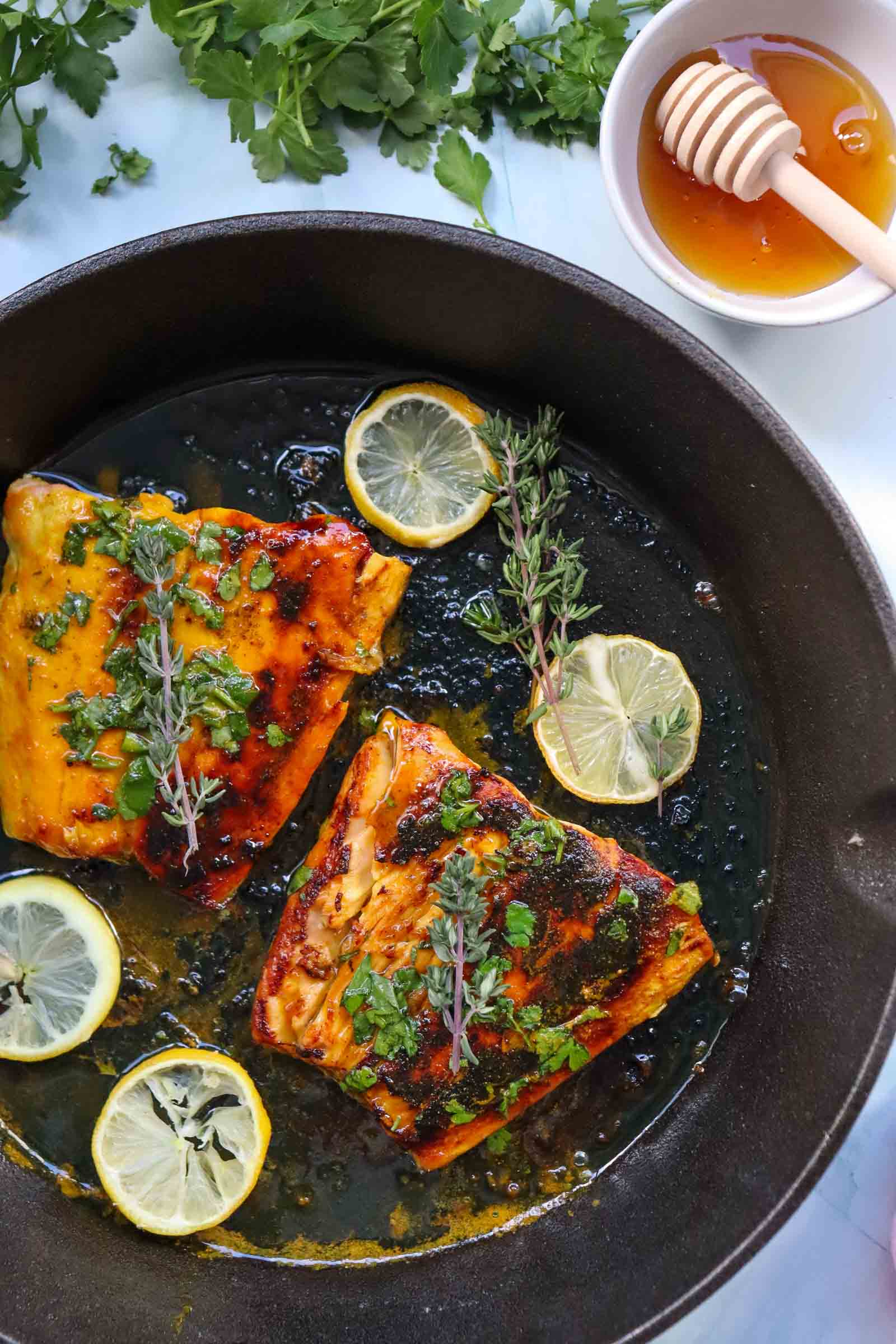 Two cooked honey glazed salmon fillets in a cast iron pan, topped with herbs and surrounded by lemon slices; honey and a dipper are in a bowl nearby.