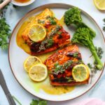 A plate with two honey glazed salmon fillets topped with lemon slices and herbs, served with broccolini. Lemon slices, fresh herbs, and a bowl of honey are nearby.
