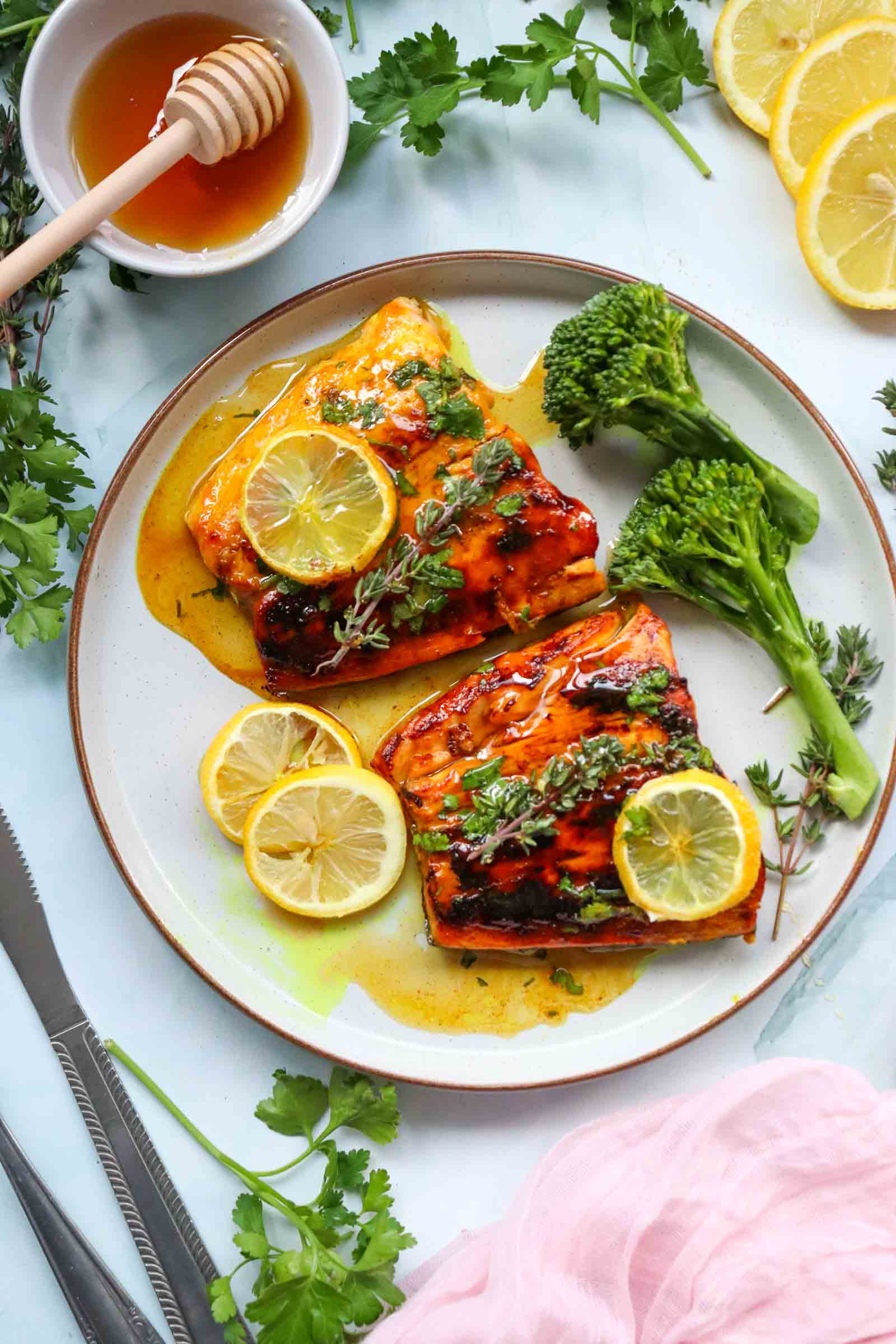 A plate with two honey glazed salmon fillets topped with lemon slices and herbs, served with broccolini. Lemon slices, fresh herbs, and a bowl of honey are nearby.