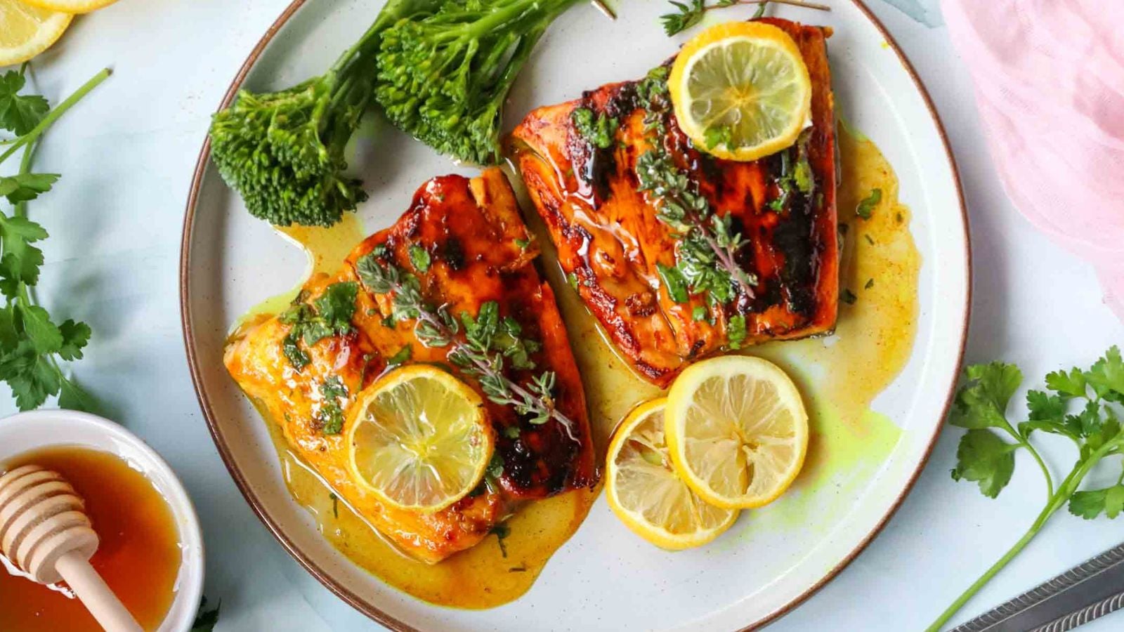 Two pieces of glazed salmon garnished with lemon slices and herbs, served on a white plate with steamed broccolini.