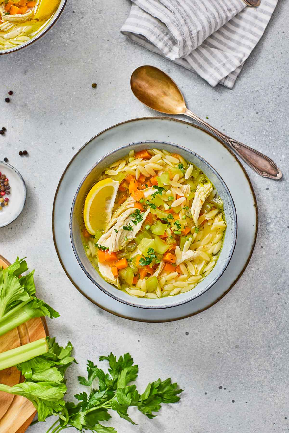 A bowl of lemon chicken orzo soup with carrots, celery, and a lemon wedge, garnished with parsley, placed on a plate with a spoon and napkin beside it.