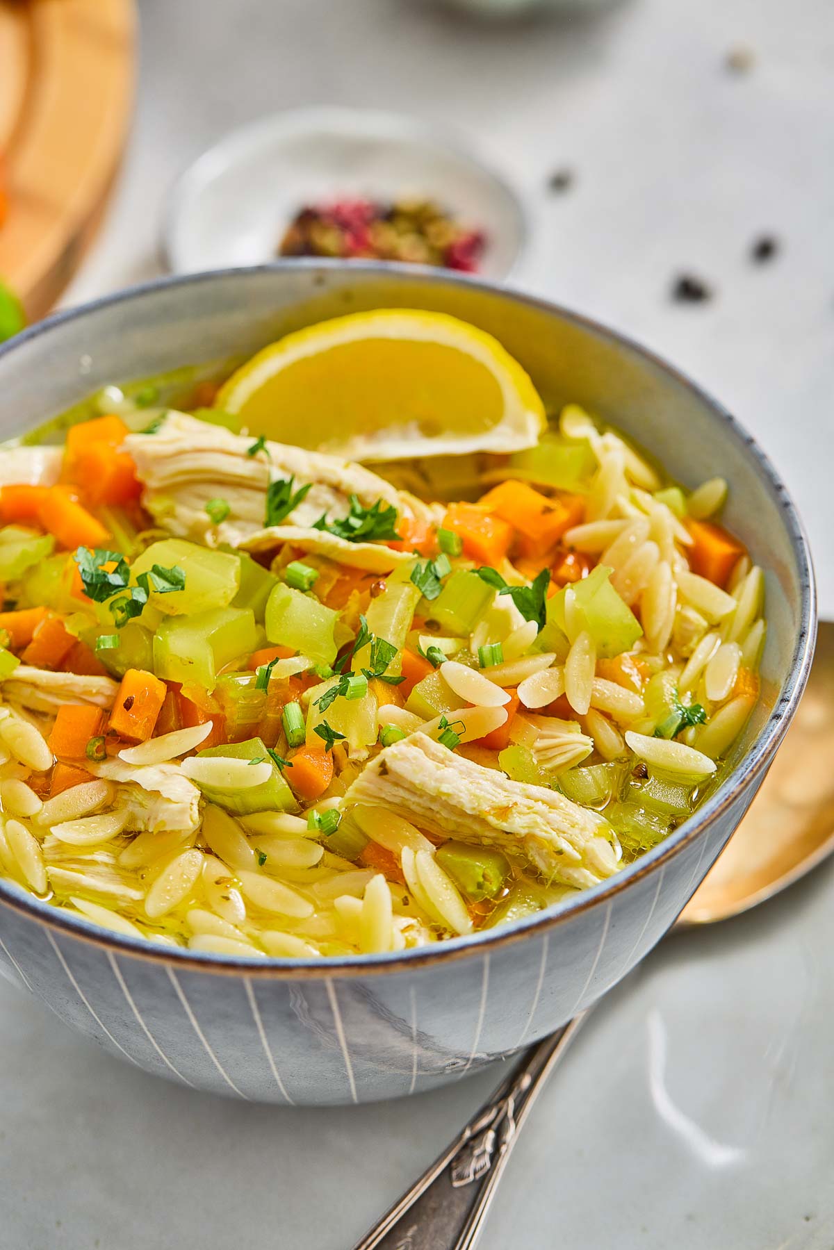 A bowl of lemon chicken orzo soup with diced carrots, celery, and parsley, garnished with a lemon wedge.