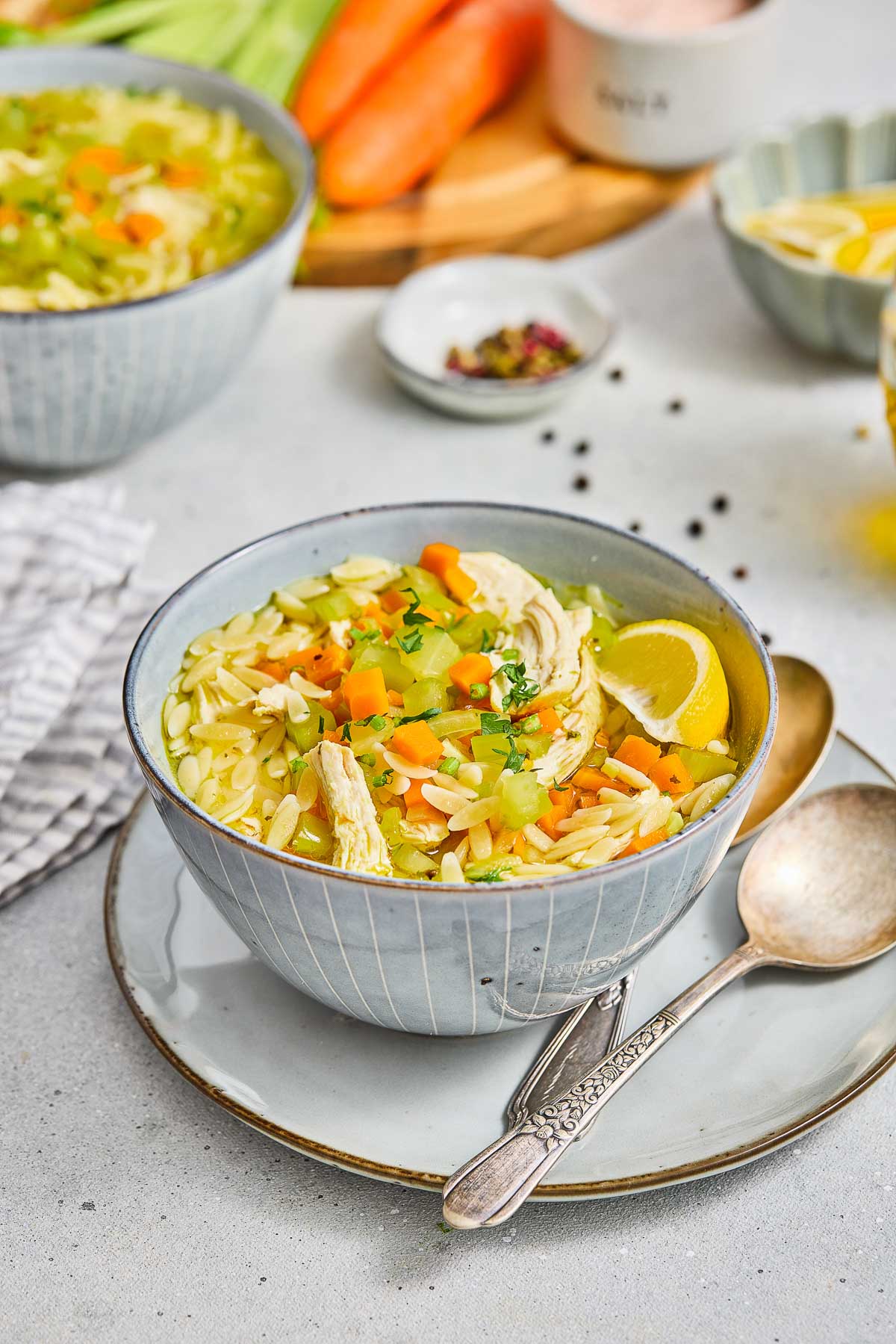 A bowl of lemon chicken orzo soup with shredded chicken, carrots, celery, orzo pasta, lemon wedge, and herbs, served with a spoon on a plate.