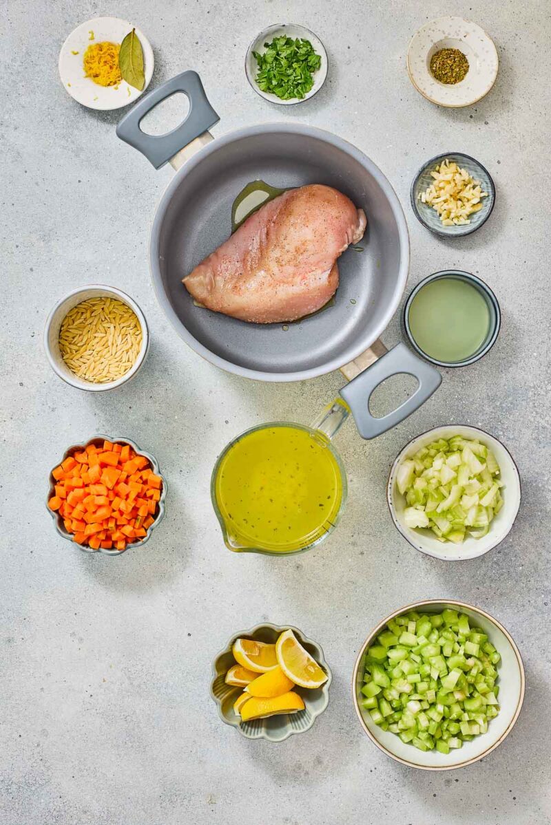 Top-down view of various ingredients for a lemon orzo chicken soup, including a raw chicken breast in a pot, chopped vegetables, lemon wedges, broth, herbs, garlic, orzo pasta, and seasonings.
