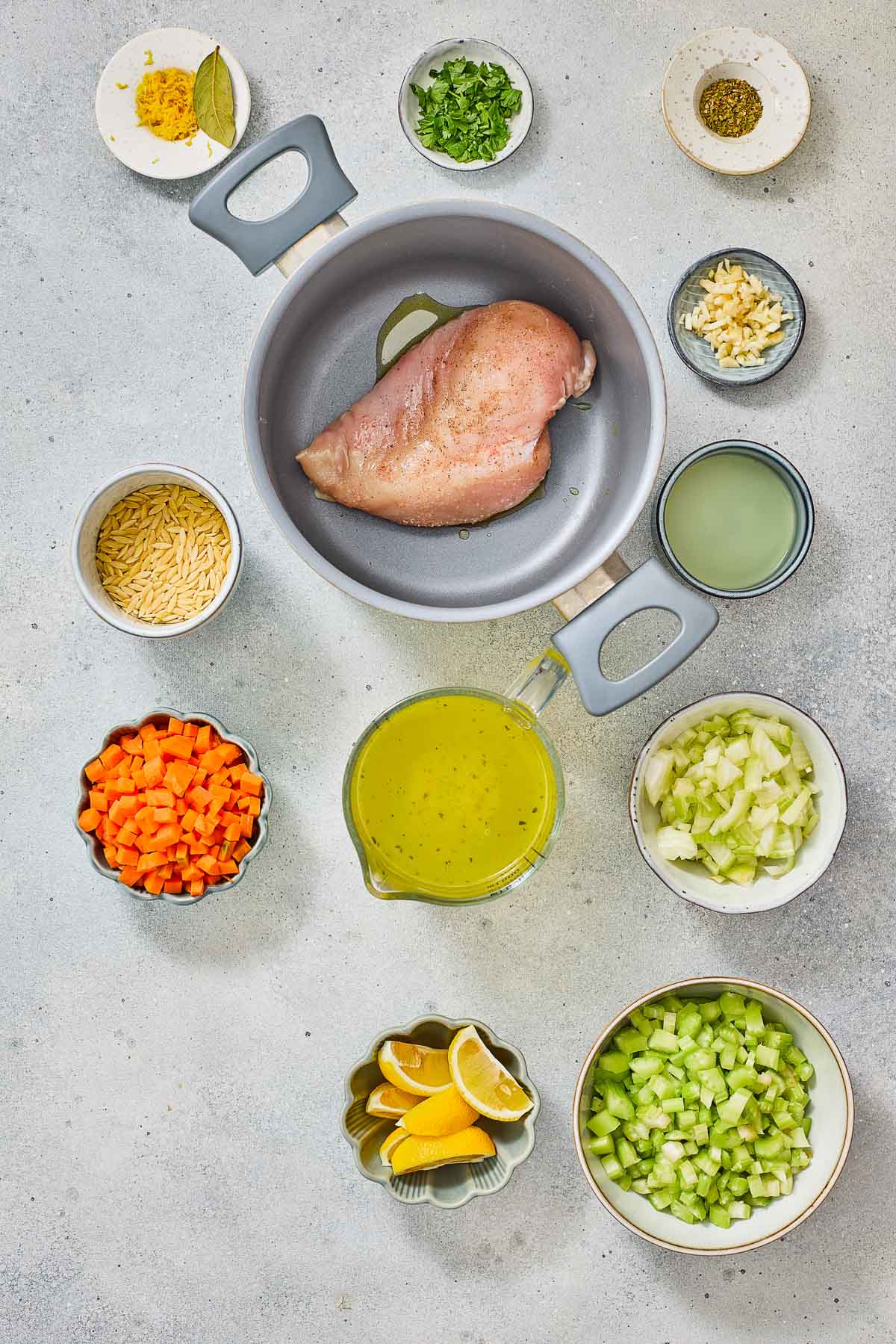 Top-down view of various ingredients for a lemon orzo chicken soup, including a raw chicken breast in a pot, chopped vegetables, lemon wedges, broth, herbs, garlic, orzo pasta, and seasonings.