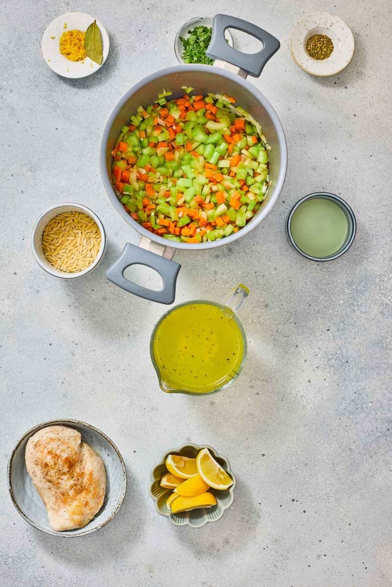 A pot with chopped carrots, celery, and onion is surrounded by cooked chicken breast, orzo, lemon wedges, broth, spices, and herbs on a light gray surface.