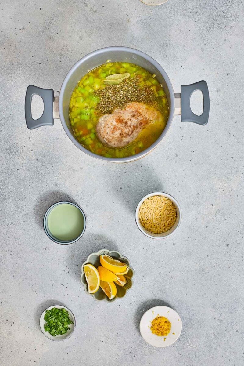 A pot with raw chicken, broth, herbs, and vegetables sits on a counter surrounded by bowls of orzo, lemon wedges, chopped herbs, and spices.