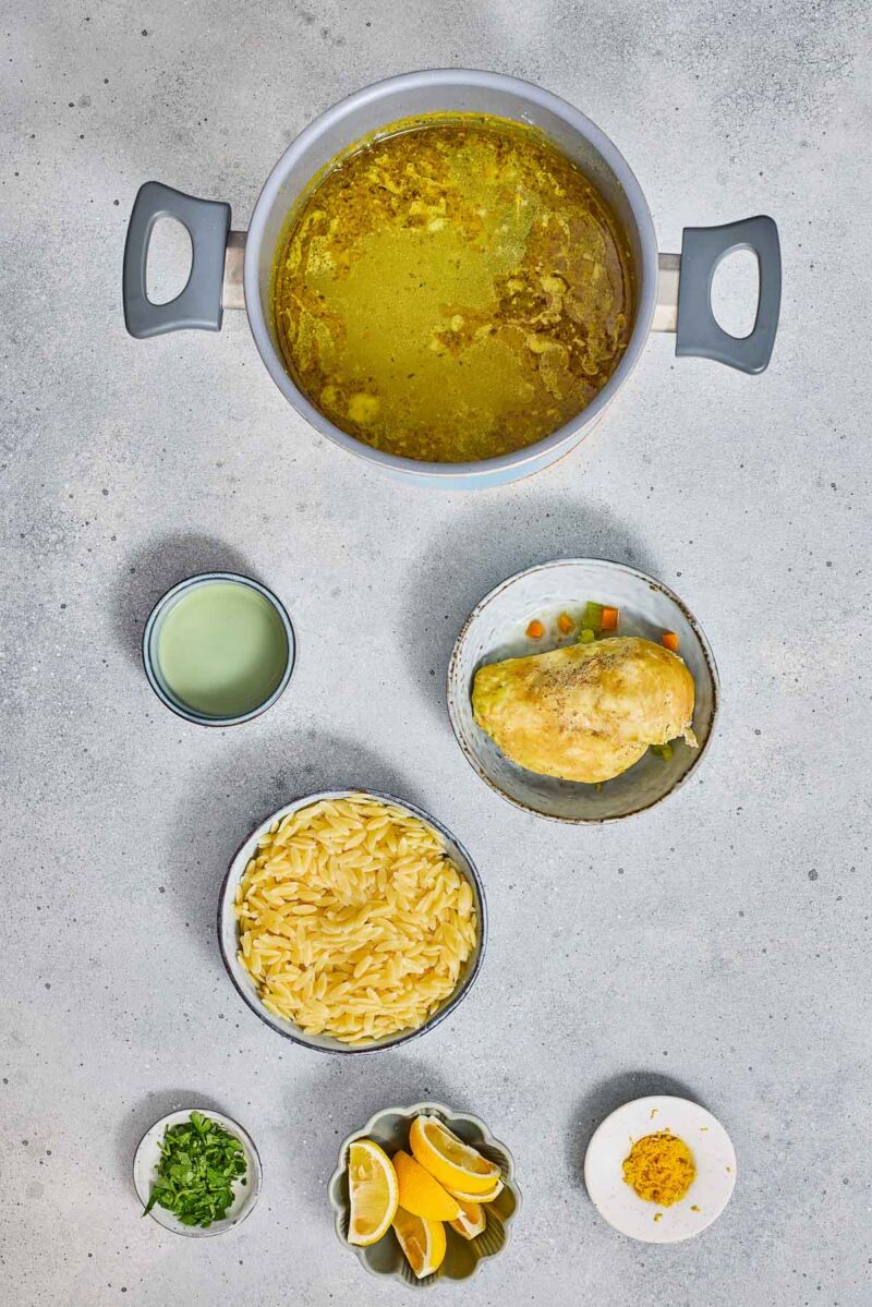Overhead view of a pot with broth, a bowl with a chicken piece and vegetables, a bowl of orzo pasta, a cup of liquid, chopped herbs, lemon wedges, and a small plate with seasoning.