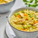 A bowl of lemon chicken soup with orzo pasta, shredded chicken, diced carrots, celery, and parsley, placed on a white plate. Another bowl is in the background.