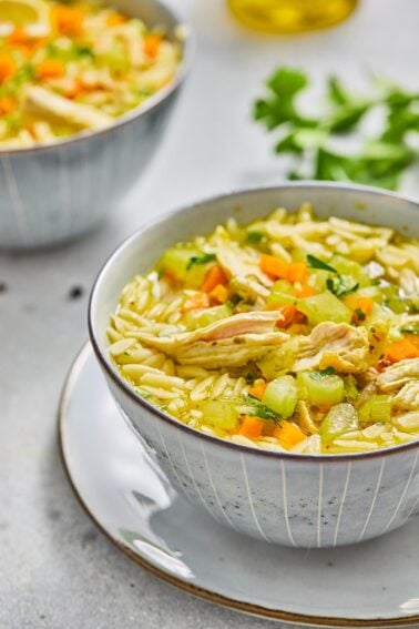 A bowl of lemon chicken soup with orzo pasta, shredded chicken, diced carrots, celery, and parsley, placed on a white plate. Another bowl is in the background.