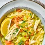 A bowl of lemon orzo chicken soup with shredded chicken, orzo, diced carrots, celery, fresh herbs, and a lemon wedge, photographed from above.