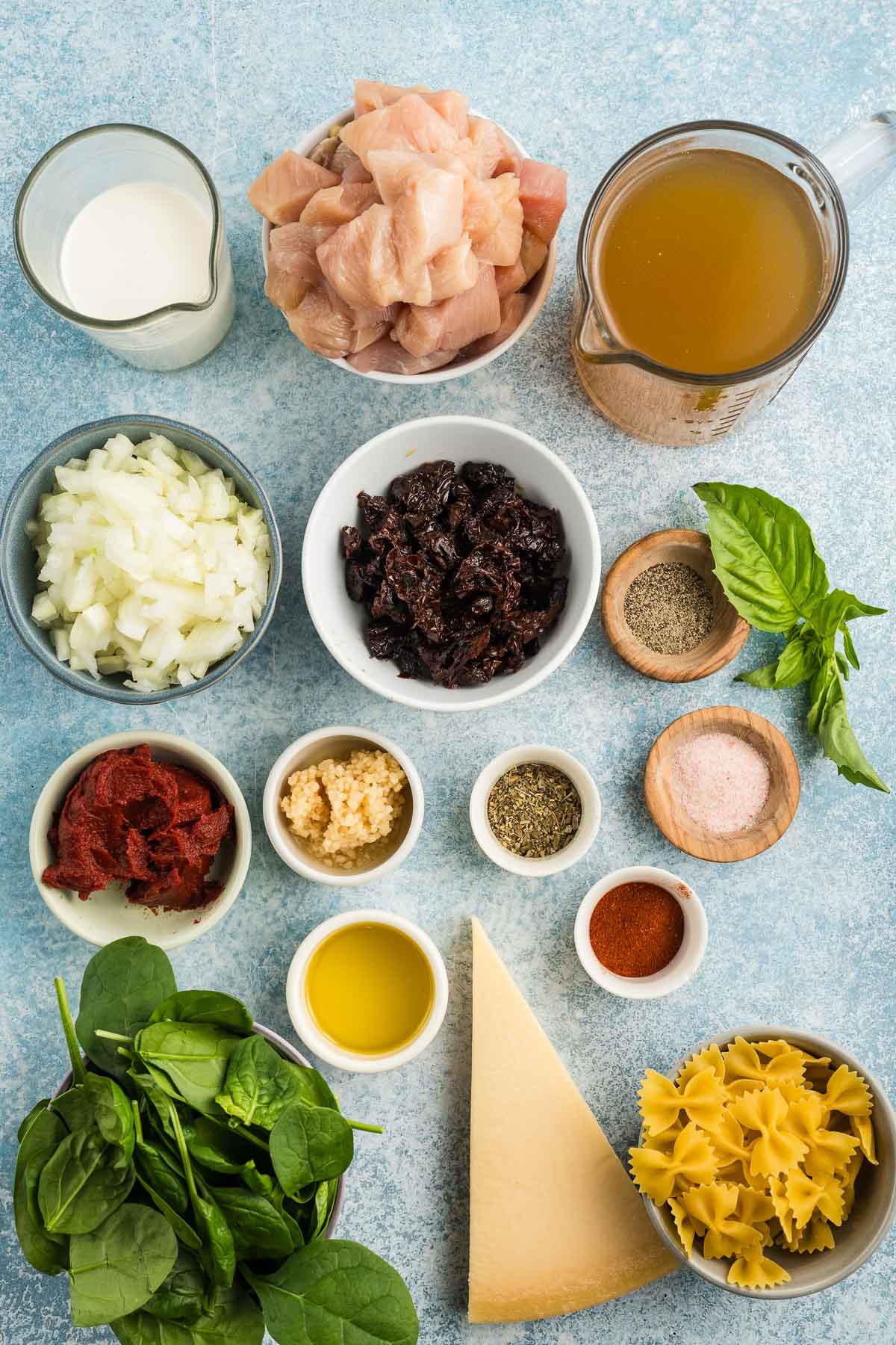 Various recipe ingredients including chopped chicken, chicken broth, heavy cream, farfalle pasta, spinach, parmesan, tomato paste, dried cherries, onion, garlic, oil, and seasonings on a counter.