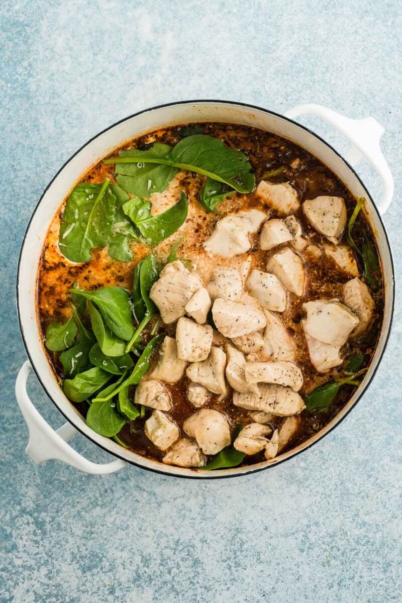 A white pot of soup filled with cooked chicken pieces, fresh spinach leaves, and broth on a light blue surface.