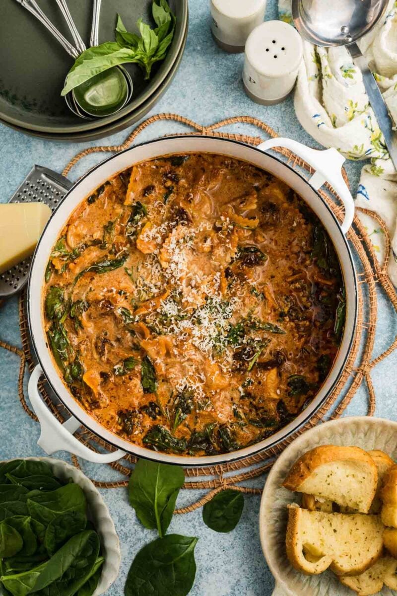 A pot of creamy marry me chicken soup garnished with grated cheese, surrounded by fresh basil, garlic bread, spinach leaves, and kitchen utensils on a blue surface.