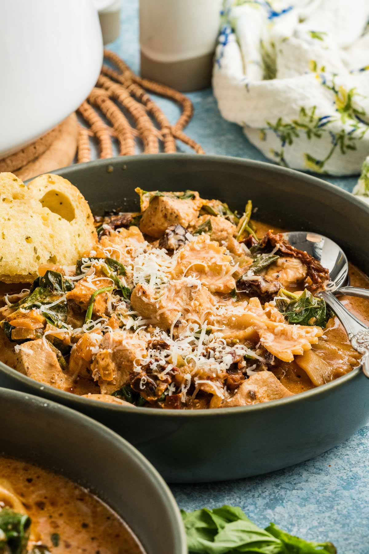 A bowl of marry me chicken soup with shredded chicken, greens, sun-dried tomatoes, pasta, grated cheese on top, and a slice of bread on the side.