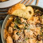 A bowl of marry me chicken soup with pasta with spinach, topped with grated cheese, served with a slice of garlic bread. A pot of the same dish is partially visible in the background.