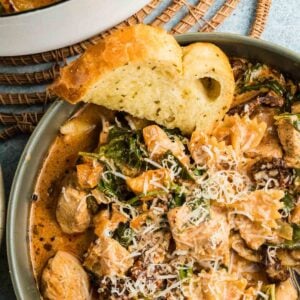 A bowl of marry me chicken soup with pasta with spinach, topped with grated cheese, served with a slice of garlic bread. A pot of the same dish is partially visible in the background.