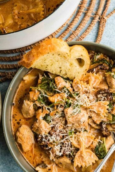 A bowl of marry me chicken soup with pasta with spinach, topped with grated cheese, served with a slice of garlic bread. A pot of the same dish is partially visible in the background.