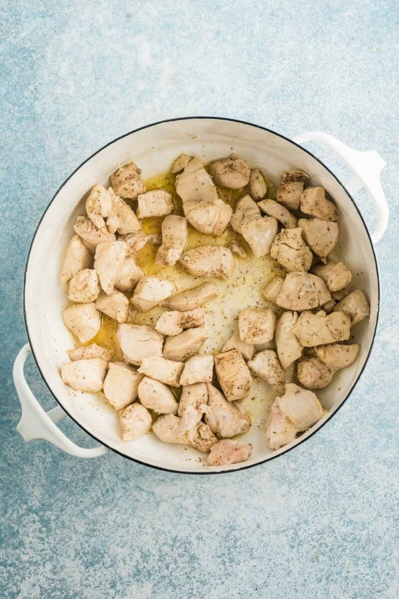 Cubed pieces of cooked chicken breast seasoned with pepper and oil in a white pot on a light blue textured surface.