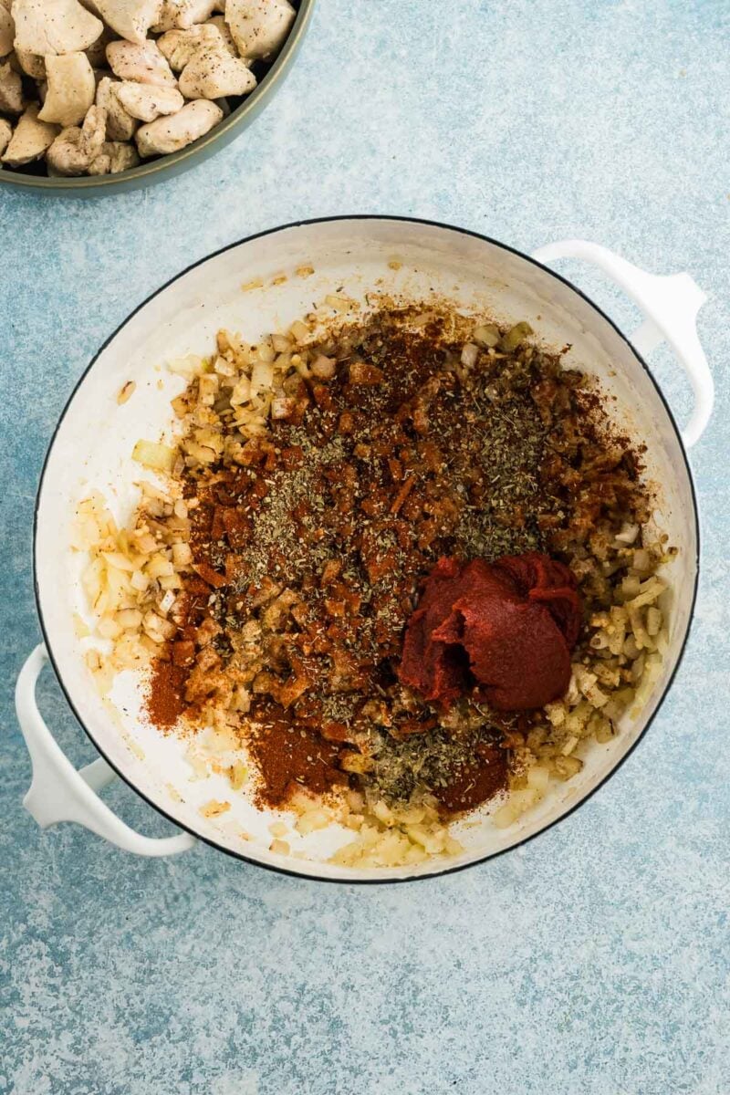 Chopped onions, herbs, spices, and tomato paste in a white pot, with a bowl of cooked chicken pieces nearby on a blue background.