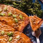 Miso glazed salmon fillets topped with sesame seeds and sliced green onions, served with broccolini on a blue plate, with a fork holding a piece of salmon.