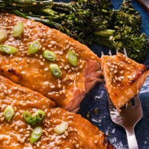 Miso glazed salmon fillets topped with sesame seeds and sliced green onions, served with broccolini on a blue plate, with a fork holding a piece of salmon.