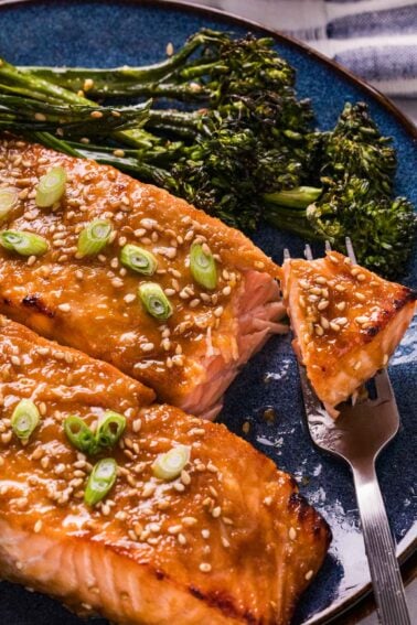 Miso glazed salmon fillets topped with sesame seeds and sliced green onions, served with broccolini on a blue plate, with a fork holding a piece of salmon.