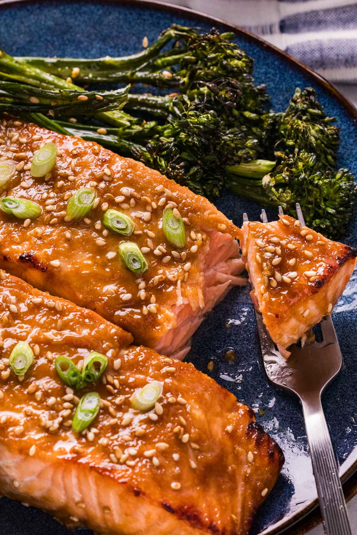 Miso glazed salmon fillets topped with sesame seeds and sliced green onions, served with broccolini on a blue plate, with a fork holding a piece of salmon.