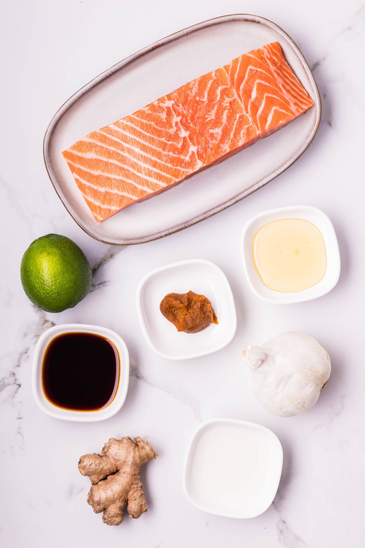 Raw salmon filet on a plate with lime, garlic, ginger, soy sauce, miso paste, honey, and a small dish of white liquid arranged on a white surface.