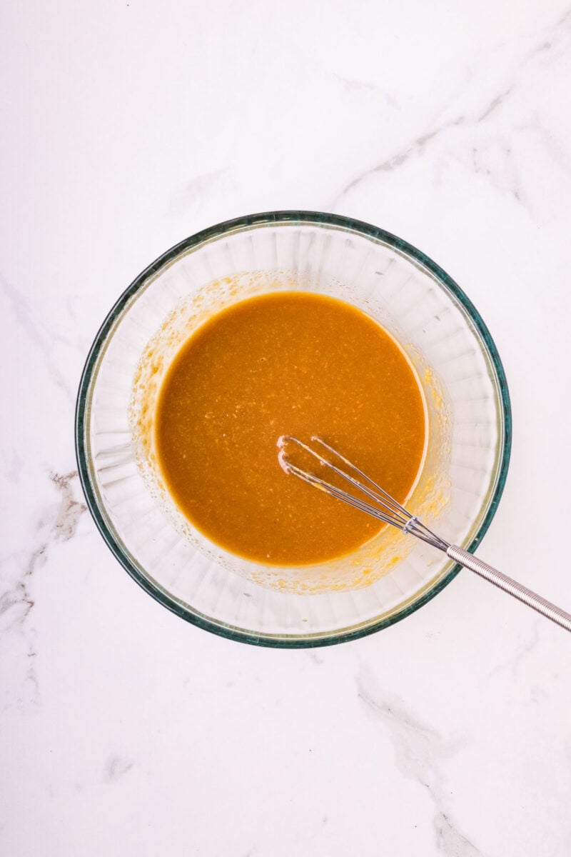 A glass bowl filled with an miso glaze mixture and a metal whisk on a white marble surface.