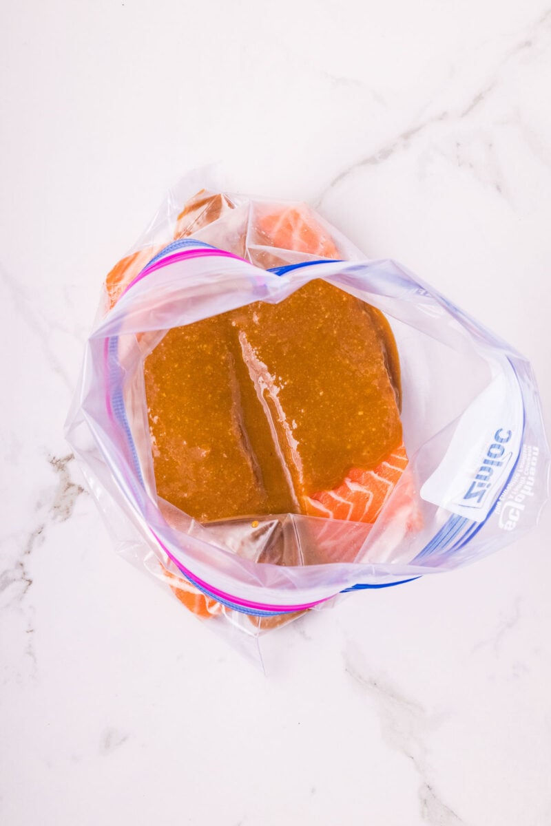 Raw salmon fillets in a resealable plastic bag with miso marinade, placed on a white marble surface.