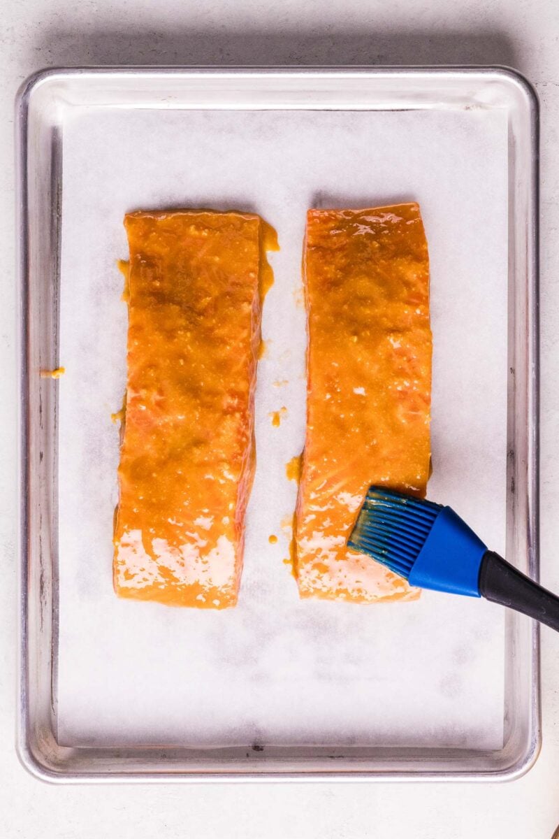 Two salmon fillets on a parchment-lined baking sheet being brushed with miso sauce using a blue silicone brush.