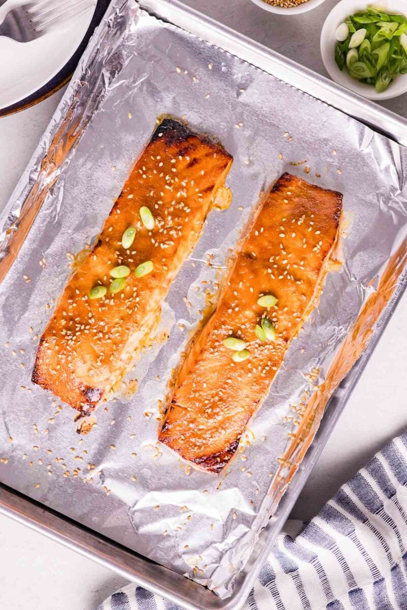 Two baked miso glazed salmon fillets on a foil-lined baking sheet, topped with sesame seeds and chopped green onions.