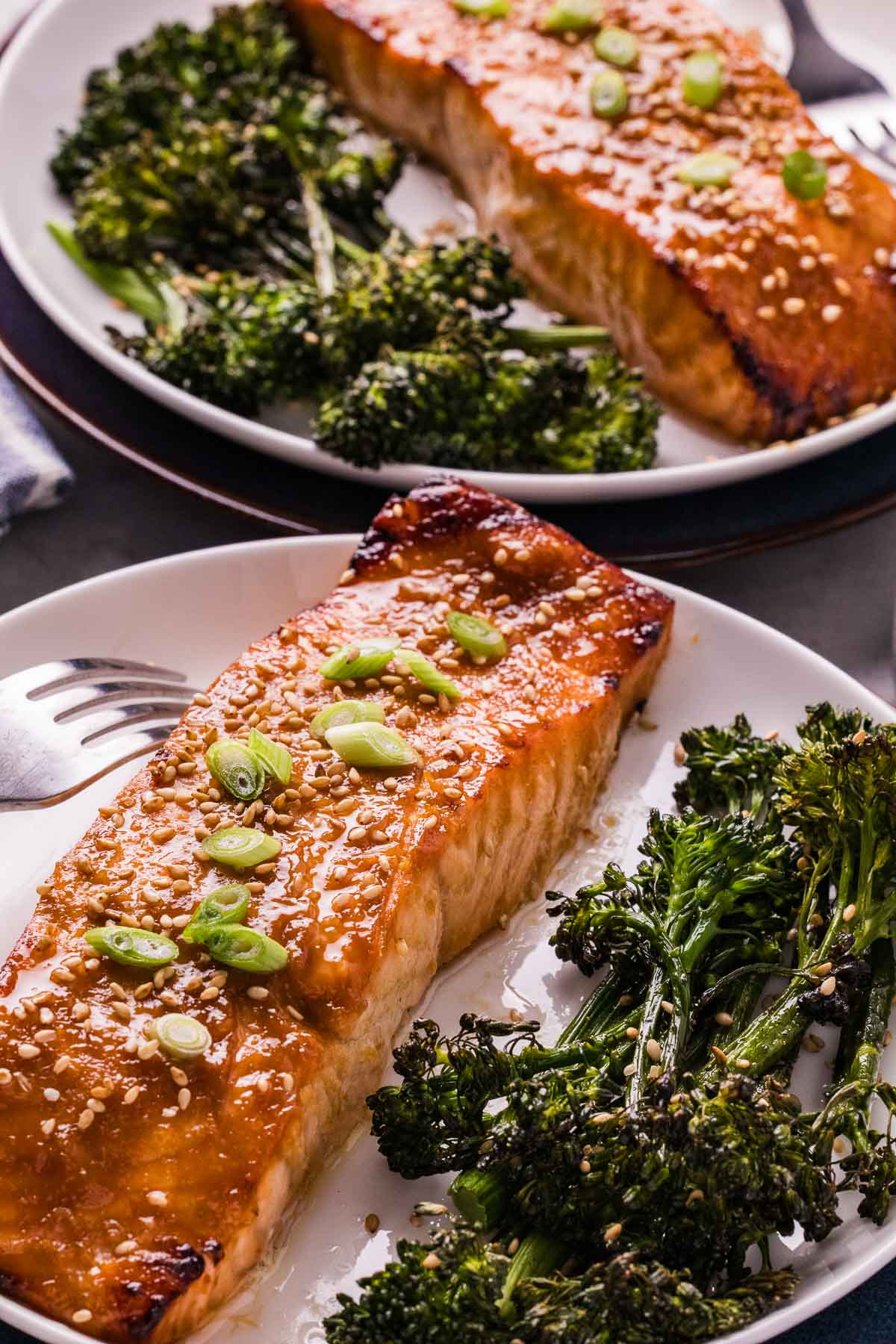 Grilled salmon fillet topped with sesame seeds and green onions, served with roasted broccolini on a white plate.