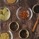 Assorted spices including bay leaves, red pepper flakes, cinnamon stick, coriander seeds, and peppercorns arranged in bowls on a wooden surface.