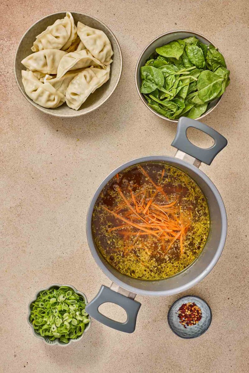 A pot of broth with shredded carrots is surrounded by bowls of dumplings, fresh spinach, chopped green onions, and red pepper flakes on a beige surface.