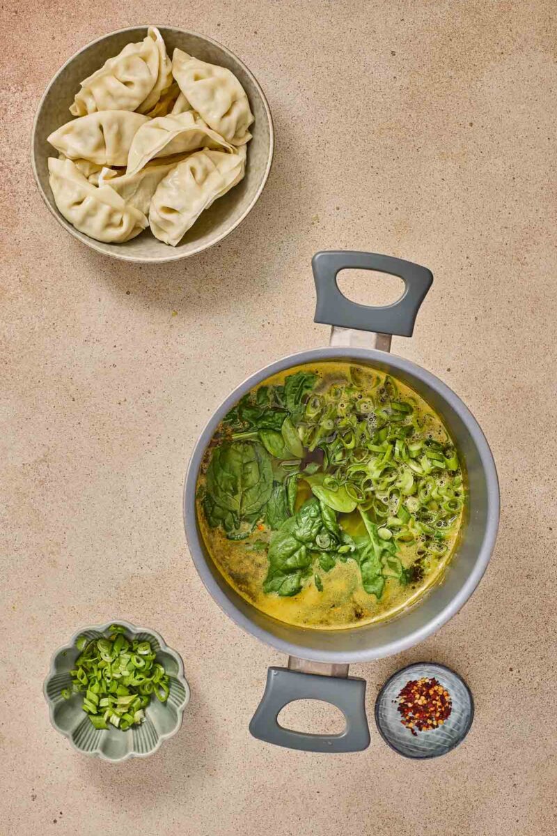 A pot of soup with spinach and scallions, a bowl of dumplings, a small dish of sliced green onions, and a small dish of red pepper flakes on a beige surface.