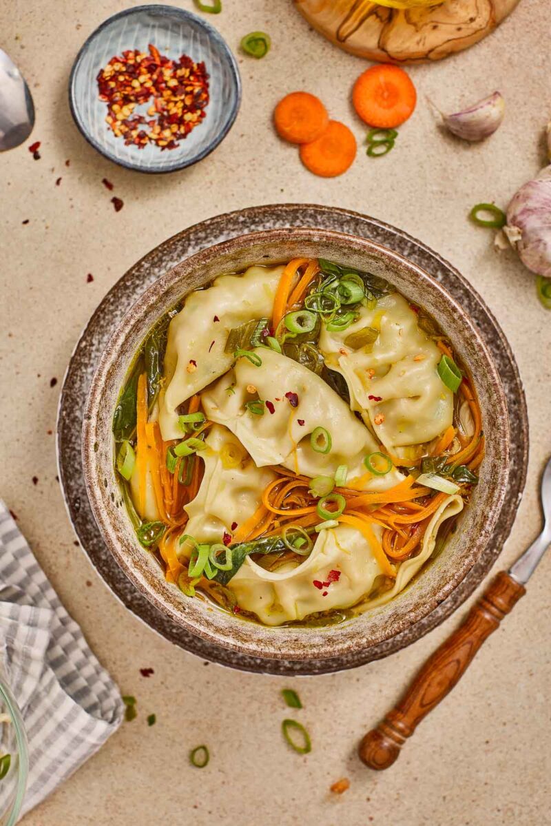 A bowl of potsticker soup with sliced carrots, green onions, and broth, surrounded by a spoon, garlic, sliced carrot, and a small dish of red pepper flakes.