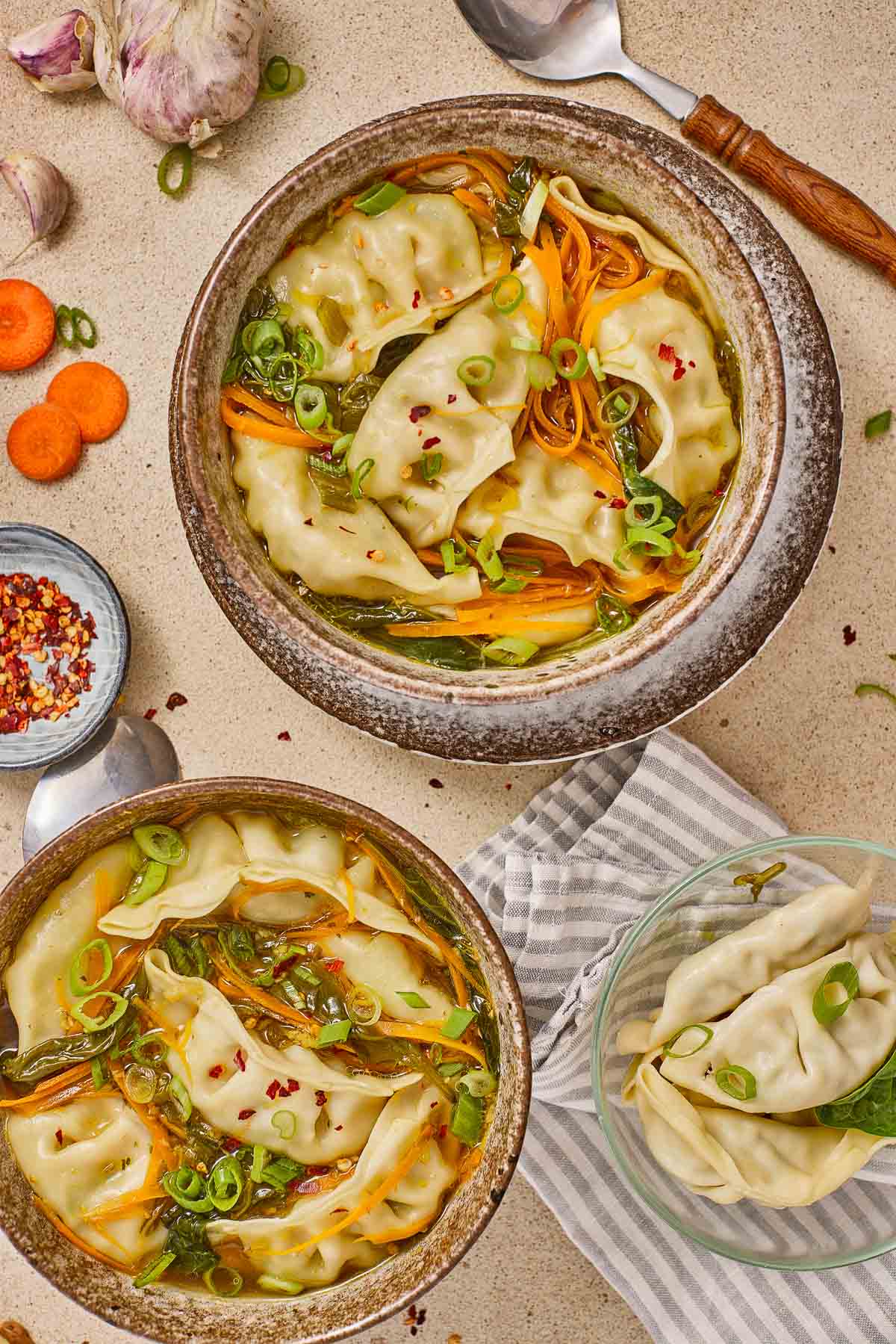 Two bowls of dumpling soup garnished with sliced green onions and vegetables, with a small bowl of dumplings on the side and spoon, garlic, and carrot slices nearby.