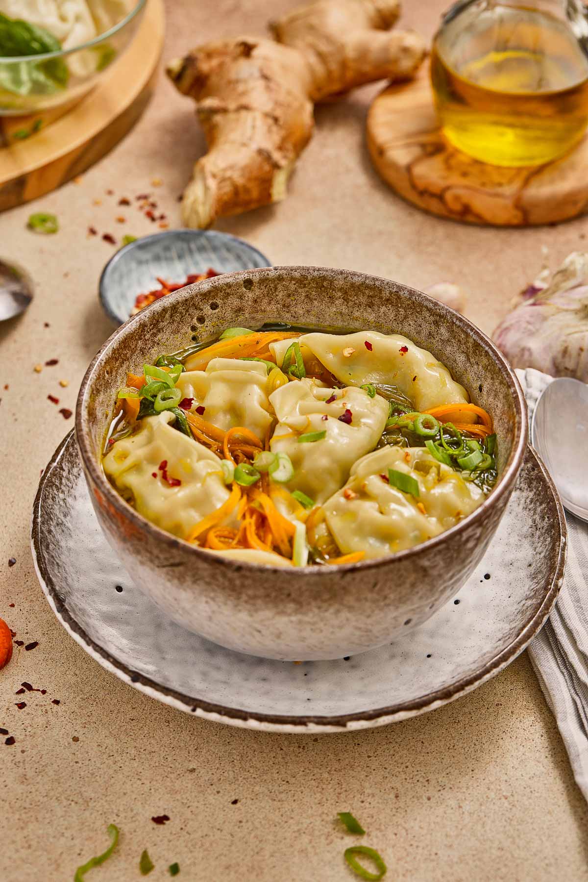 A bowl of potsticker soup garnished with sliced green onions and shredded carrots, with ginger, oil, and other ingredients in the background.