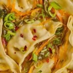 Close-up of dumplings in a soup broth with sliced green onions, shredded carrots, leafy greens, and red pepper flakes.