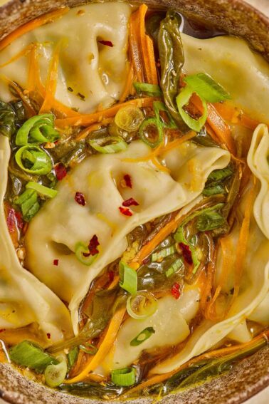 Close-up of dumplings in a soup broth with sliced green onions, shredded carrots, leafy greens, and red pepper flakes.