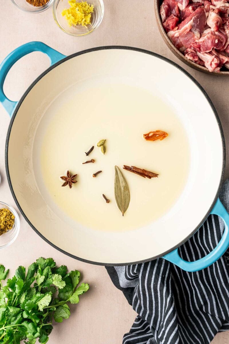 A large pot with oil and whole spices including bay leaf, cinnamon stick, star anise, cloves, cardamom, and dried chili, surrounded by raw meat, cilantro, and small bowls of ingredients.