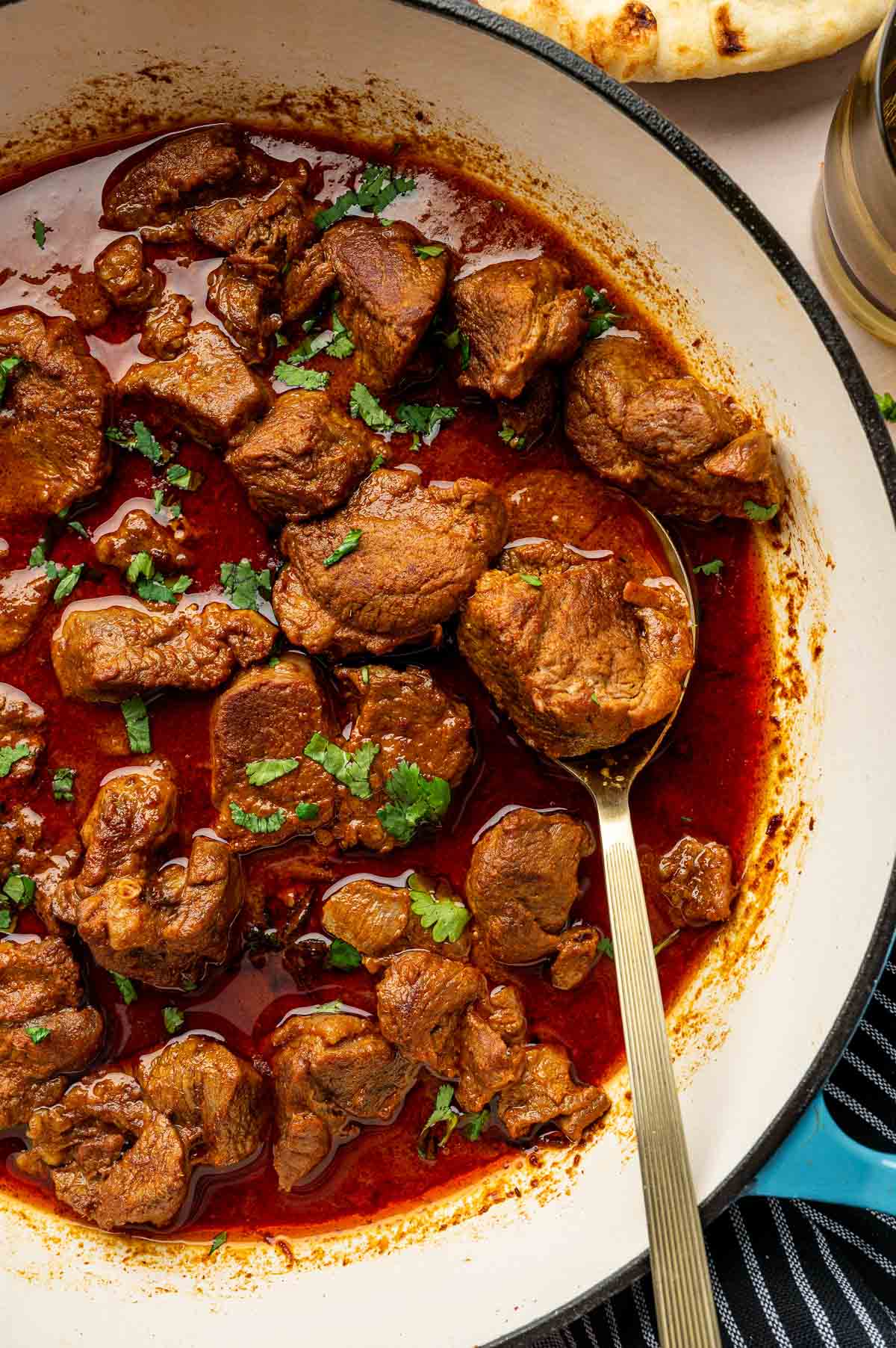 A pot of rogan josh in a rich red sauce, garnished with chopped herbs, with a spoon in the dish.