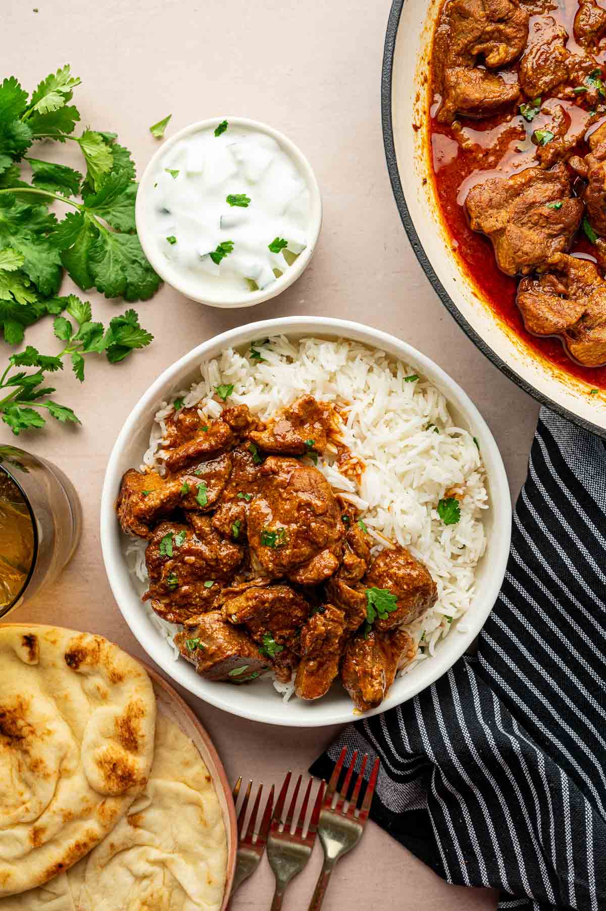A bowl of white rice topped with kashmiri lamb curry, served with naan, a glass of water, a bowl of raita, fresh cilantro, and a striped napkin.