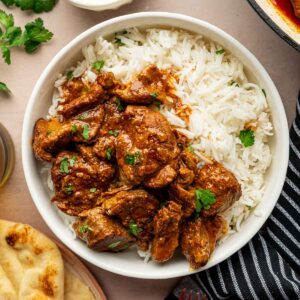 A bowl of white rice topped with rogan josh, garnished with cilantro, served with naan, yogurt, and fresh cilantro on the side.